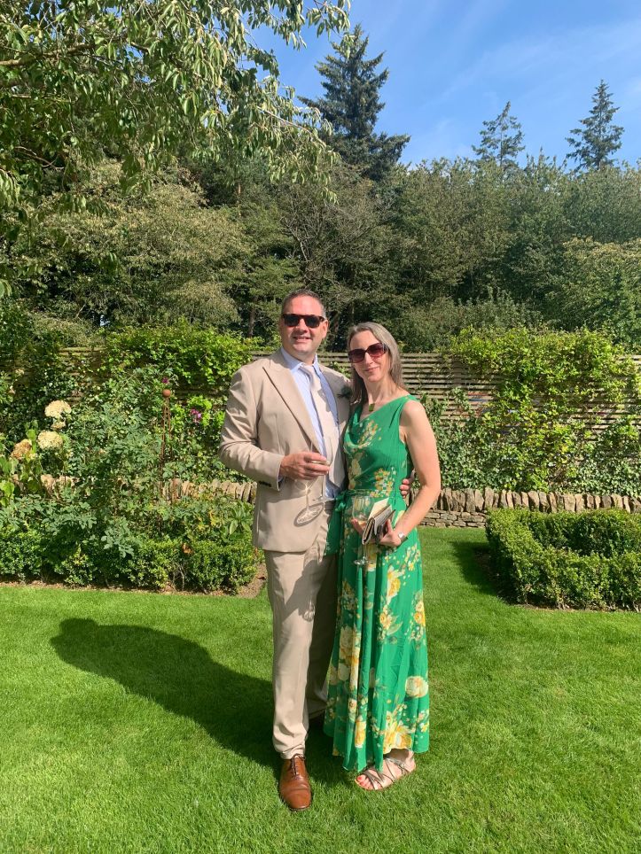 Photo of a couple in a garden. The man is wearing a stone coloured suit, brown shoes and sunglasses. The woman is wearing a bright green dress, silver sandals and sunglasses.